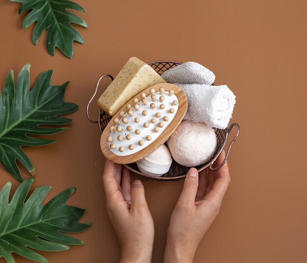 Nature morte avec des mains féminines, brosse de massage, bombes de bain, savon et une serviette dans un panier avec vue de dessus de feuilles.