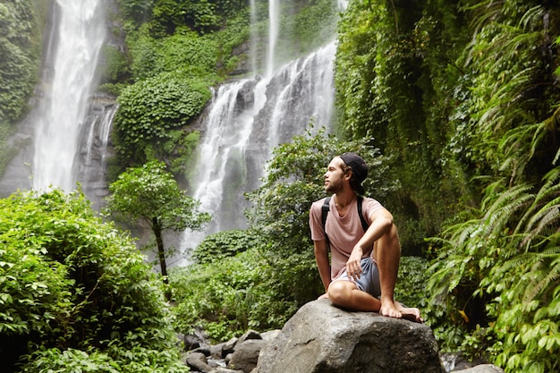 Nature, tourisme et hommes. Jeune touriste pieds nus portant des jeans courts et sac à dos assis sur un gros rocher au fond de la forêt tropicale verte