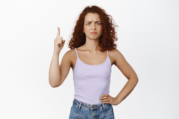 Photo gratuite ne pas aimer. portrait de jeune femme rousse bouclée fronçant les sourcils, pointant le doigt vers le haut et fronçant les sourcils mécontent, n'aimant pas qch, debout sur fond blanc