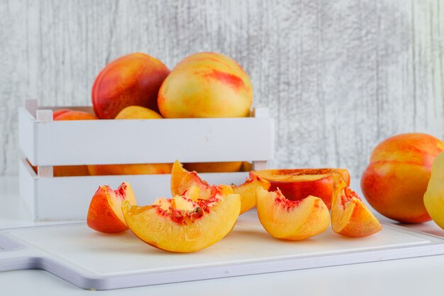 Nectarines avec planche à découper dans une boîte en bois sur mur blanc et grungy, vue latérale.