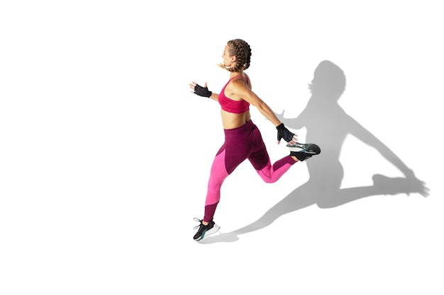 Énergie. Belle jeune athlète féminine pratiquant sur un mur blanc, portrait avec des ombres. Modèle de coupe sportive en mouvement et en action. Musculation, mode de vie sain, concept de style.