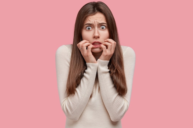 Photo gratuite nerveuse belle femme a un regard inquiet, se sent perplexe et stressée avant la session d'examen, garde les mains près de la bouche, vêtue d'un pull blanc