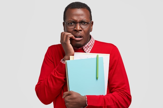 Nerveux anxieux homme à la peau sombre mord les doigts, regarde avec insistance, tient des manuels, fronce les sourcils de mécontentement, porte un pull rouge, une chemise élégante