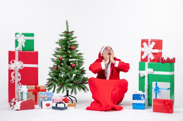 Nerveux jeune adulte émotionnel habillé en père Noël avec des cadeaux et arbre de Noël décoré assis sur le sol à la recherche au-dessus sur fond blanc