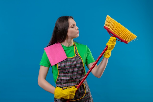 Nettoyage jeune femme en uniforme dans des gants à la recherche de vadrouille sur sa main sur un mur bleu isolé