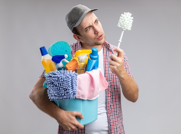 nettoyeur de jeune homme confus portant une casquette tenant un seau avec des outils de nettoyage et regardant une brosse dans sa main isolée sur un mur blanc