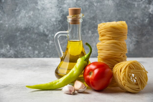 Nids de pâtes sèches, bouteille d'huile d'olive et légumes sur tableau blanc.