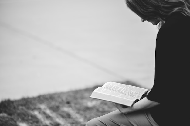 Photo gratuite niveaux de gris d'une femme assise à l'extérieur en lisant un livre