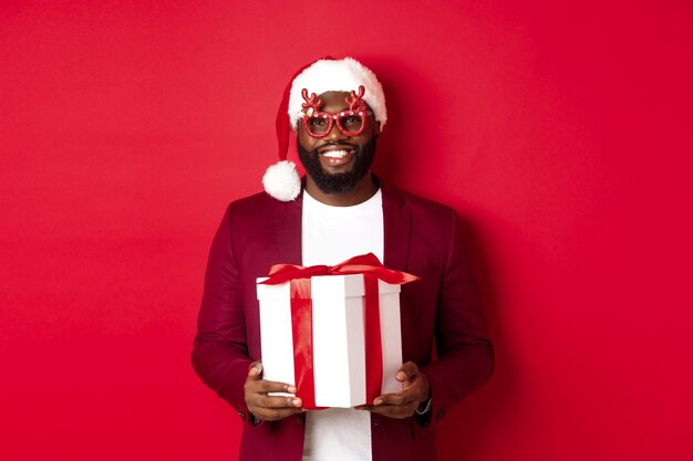 Noël. Bel homme afro-américain dans des verres de fête et bonnet de Noel tenant le cadeau du nouvel an