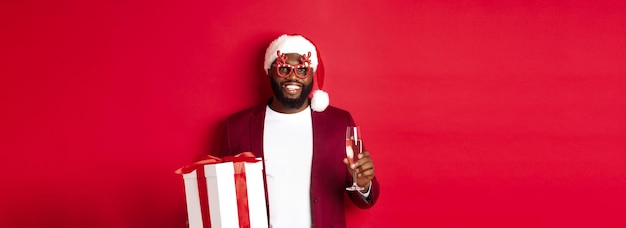 Photo gratuite noël bel homme afro-américain dans des verres de fête et bonnet de noel tenant un cadeau de nouvel an et gla