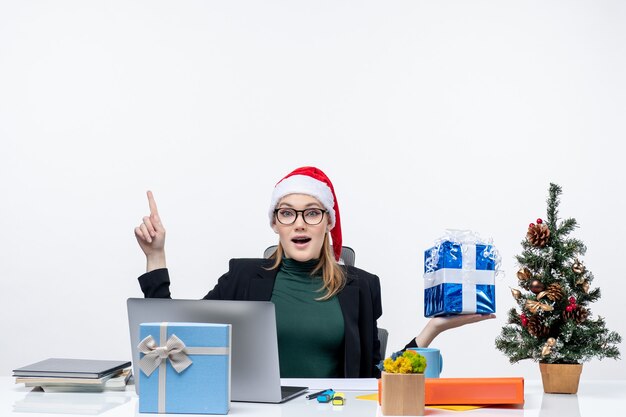 Noël - humeur festive avec curieuse jeune femme positive avec chapeau de père Noël et portant des lunettes assis à une table montrant un cadeau pointant au-dessus sur fond blanc