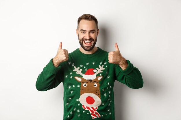 Noël, vacances et célébration. Heureux jeune homme appréciant la fête du Nouvel An, montrant les pouces vers le haut en signe d'approbation, comme quelque chose de bien, debout sur fond blanc