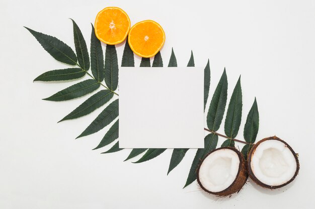 Noix de coco coupées en deux; oranges juteuses et papier blanc sur une feuille verte sur fond blanc