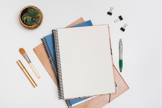 Note en spirale vierge avec un pinceau de maquillage; mascara; trombones et stylo avec des plantes en pot sur le bureau blanc