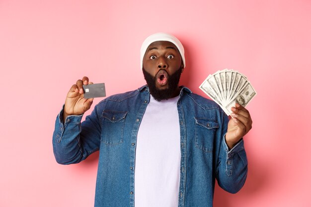 Notion de magasinage. Homme afro-américain excité montrant une carte de crédit et des dollars, a obtenu un dépôt ou un prêt d'argent, debout sur fond rose
