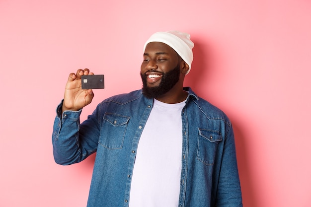 Notion de magasinage. Image d'un homme afro-américain heureux regardant satisfait de la carte de crédit, recommandant la banque, debout sur fond rose.