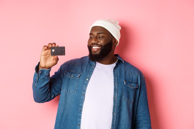 Notion de magasinage. Image d'un homme afro-américain heureux regardant satisfait de la carte de crédit, recommandant la banque, debout sur fond rose.