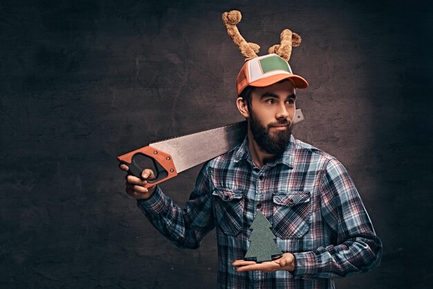 Notion de Noël. Ouvrier, charpentier, homme à tout faire portant une chemise à carreaux et une casquette de noël décorée pose avec une scie.