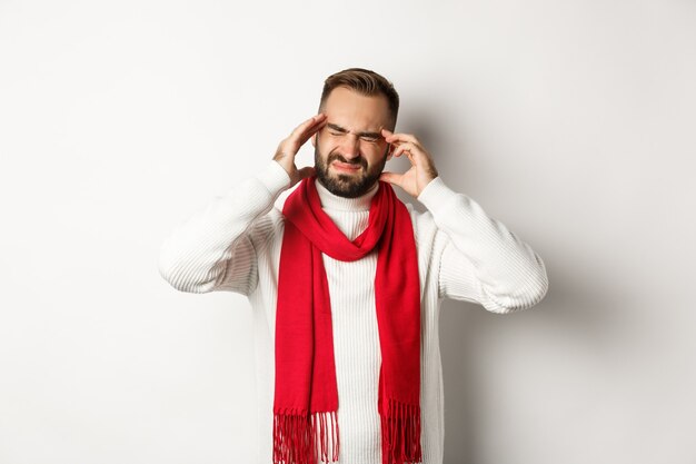 Photo gratuite notion de santé. homme ayant de graves maux de tête, touchant la tête et grimaçant de migraine, debout dans un pull d'hiver et une écharpe rouge, fond blanc