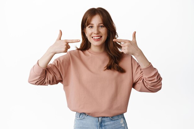 Notion de stomatologie. Femme rousse souriante pointant du doigt son sourire blanc, montrant les dents après le rendez-vous chez le dentiste, debout sur fond blanc