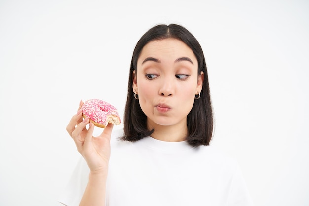 Photo gratuite nourriture et manger à l'extérieur jeune femme coréenne regarde prend une bouchée de délicieux beignet glacé rose dos blanc