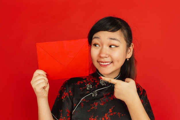 Nouvel an chinois 2020. Portrait de jeune fille asiatique isolé sur fond rouge. Gros plan du modèle féminin en vêtements traditionnels semble heureux et montrant une enveloppe rouge. Célébration, vacances, émotions.