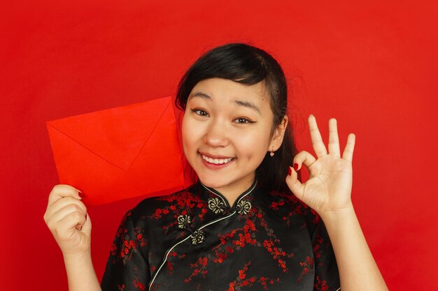 Nouvel An chinois. Portrait de jeune fille asiatique isolé sur fond rouge. Gros plan du modèle féminin en vêtements traditionnels semble heureux et montrant une enveloppe rouge. Célébration, vacances, émotions.