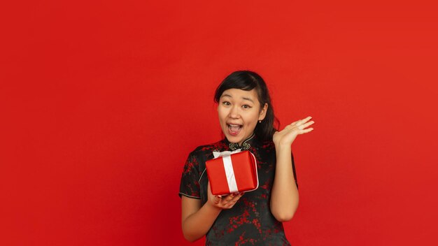 Nouvel An chinois. Portrait de jeune fille asiatique isolé sur fond rouge. Le modèle féminin en vêtements traditionnels a l'air heureux, souriant et surpris par le coffret. Célébration, vacances, émotions.