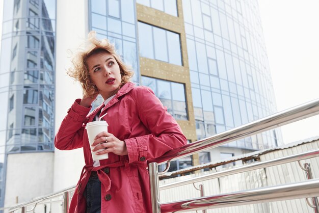 Nouvelle boisson fraîche. Jolie femme adulte en manteau rouge chaud se promène dans la ville à son heure du week-end