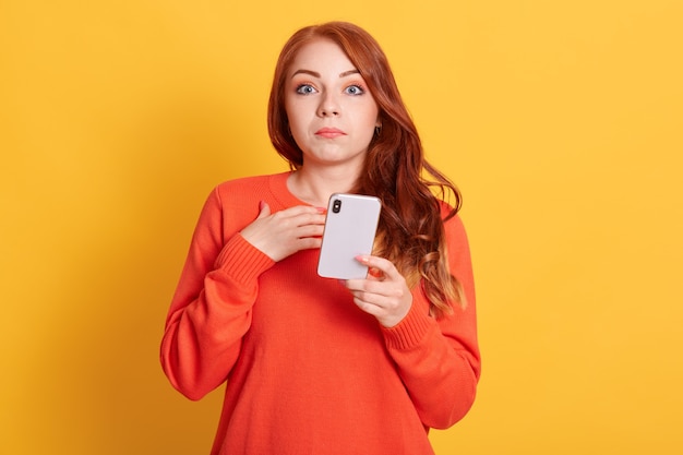 Des nouvelles choquantes! Bouchent le portrait d'une jeune femme surprise en pull décontracté orange, lecture de nouvelles sensationnelles sur téléphone intelligent