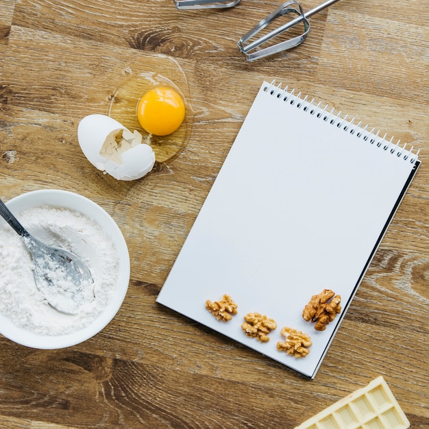 Noyer; bloc-notes en spirale; farine; chocolat et fouet sur une surface en bois