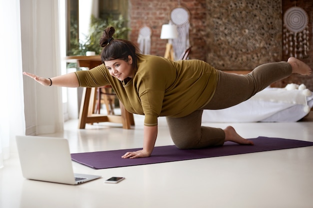 Obèse potelée jeune femme européenne avec noeud de cheveux pratiquant le yoga ou le pilates à l'intérieur sur un tapis, faisant des exercices pour renforcer le tronc, regardant une leçon vidéo en ligne devant un ordinateur portable ouvert sur le sol