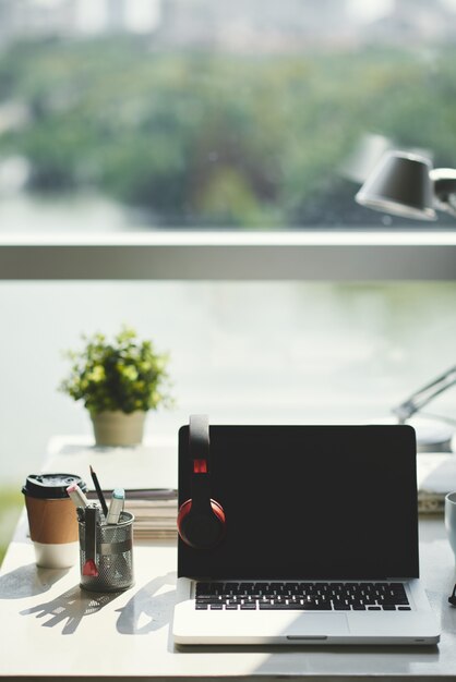 Objet: ordinateur portable éteint avec capot ouvert, écran noir et écouteurs, sur le bureau dans le bureau devant la fenêtre pendant la journée