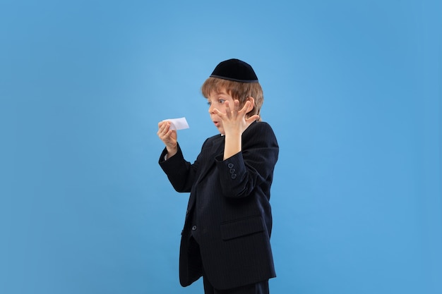 Obtenir de l'argent. Portrait d'un jeune garçon juif orthodoxe isolé sur le mur bleu du studio.