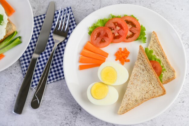 Oeufs durs, pain, carottes et tomates sur une plaque blanche avec un couteau et une fourchette.