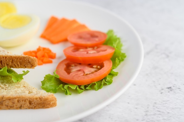 Oeufs durs, pain, carottes et tomates sur une plaque blanche avec un couteau et une fourchette.