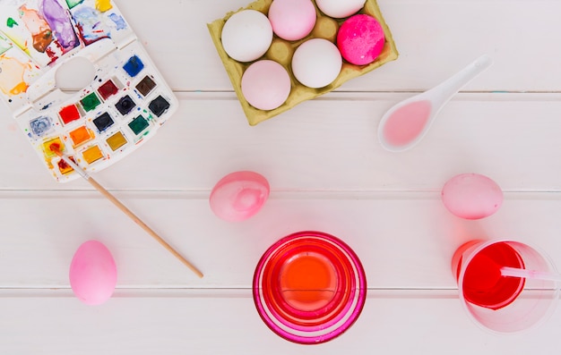 Photo gratuite oeufs de pâques dans un récipient entre des tasses de liquide de teinture près de cuillères et un pinceau avec un ensemble de couleurs