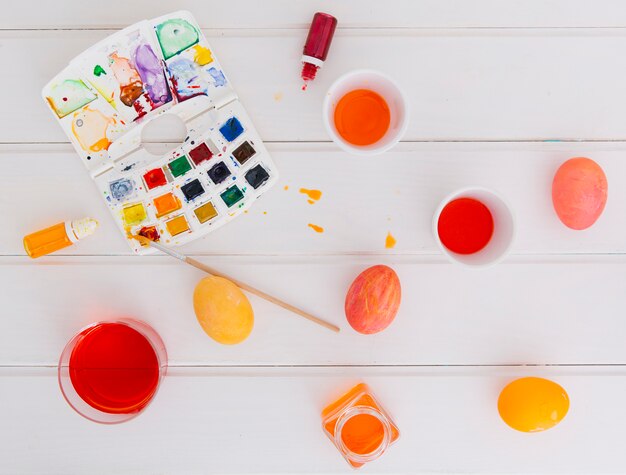 Oeufs de Pâques entre les tasses de liquide de teinture près de pinceau avec jeu de couleurs