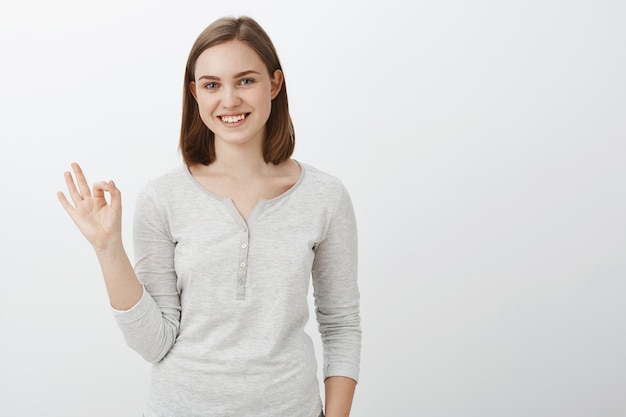 OK. Sympathique jeune femme heureuse et créative avec une coupe de cheveux brune courte montrant signe ok ou parfait avec bras levé souriant joyeusement assurant qu'elle fera le travail à temps sur un mur gris