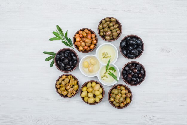 Olives colorées et huile d'olive avec des feuilles d'olivier dans une argile et des bols blancs sur planche de bois blanc, vue de dessus.