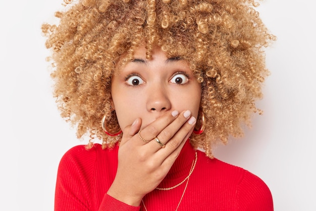 Omg concept Une belle jeune femme aux cheveux bouclés couvre la bouche avec les mains regarde les yeux sur écoute se tient sans voix porte un col roulé rouge réagit sur quelque chose d'étonnant isolé sur fond blanc