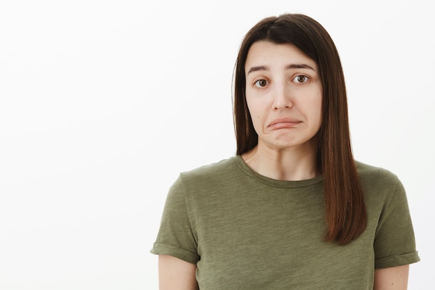 Oops Désolé. Portrait De L'insécurité Coupable Et Timide Jolie Brune Mignonne Et Le Menton Vacillant Des Lèvres Inférieures, Faisant Hausser Les épaules Comme Faisant Une Erreur, Posant Triste Et Hésitant Contre Le Mur Gris, Criant à