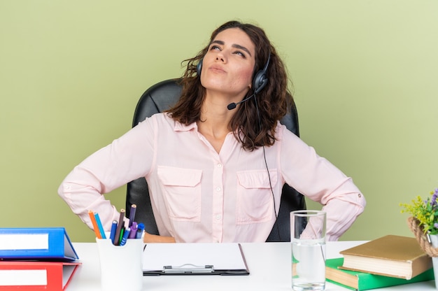 Opérateur de centre d'appels féminin assez caucasien réfléchi sur des écouteurs assis au bureau avec des outils de bureau en levant