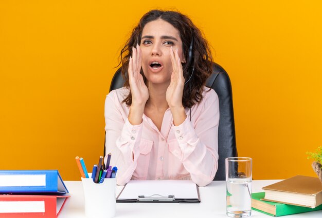 Opérateur de centre d'appels féminin assez choqué sur des écouteurs assis au bureau avec des outils de bureau en gardant les mains près de sa bouche