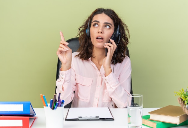 Opérateur de centre d'appels féminin assez surpris sur un casque assis au bureau avec des outils de bureau parlant au téléphone regardant et pointant vers le haut isolé sur un mur vert