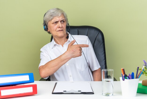 Opérateur de centre d'appels féminin caucasien mécontent sur un casque assis au bureau avec des outils de bureau regardant et pointant sur le côté isolé sur un mur vert