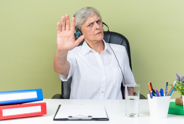 Opérateur de centre d'appels féminin caucasien sérieux sur des écouteurs assis au bureau avec des outils de bureau faisant des gestes signe de la main d'arrêt isolé sur mur vert