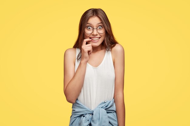 Optimiste jeune brune avec des lunettes posant contre le mur jaune