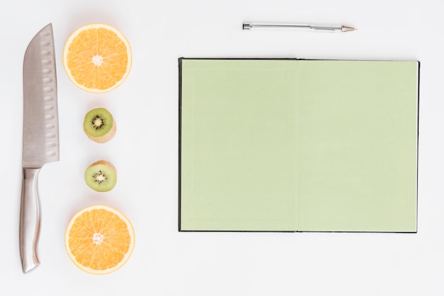 Des oranges coupées en deux; kiwi; couteau; stylo et cahier de page vierge sur fond blanc