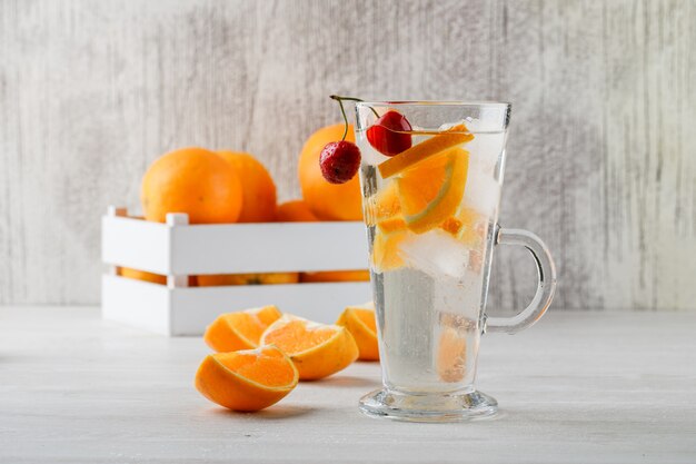 Oranges dans une boîte en bois avec vue de côté de l'eau infusée de fruits sur la surface blanche